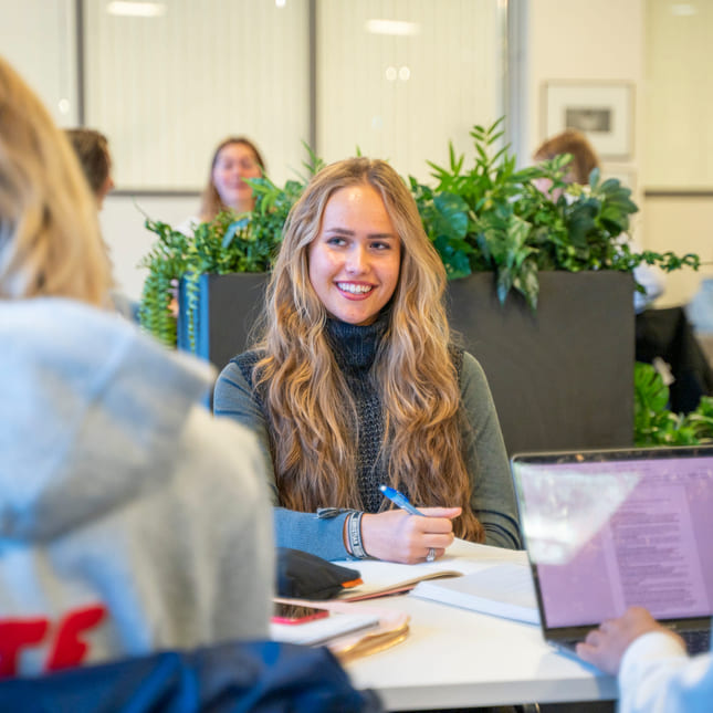 Elev smiler til medelev mens hun jobber med skole i veiledningstid på Akademiet VGS Kristiansand.