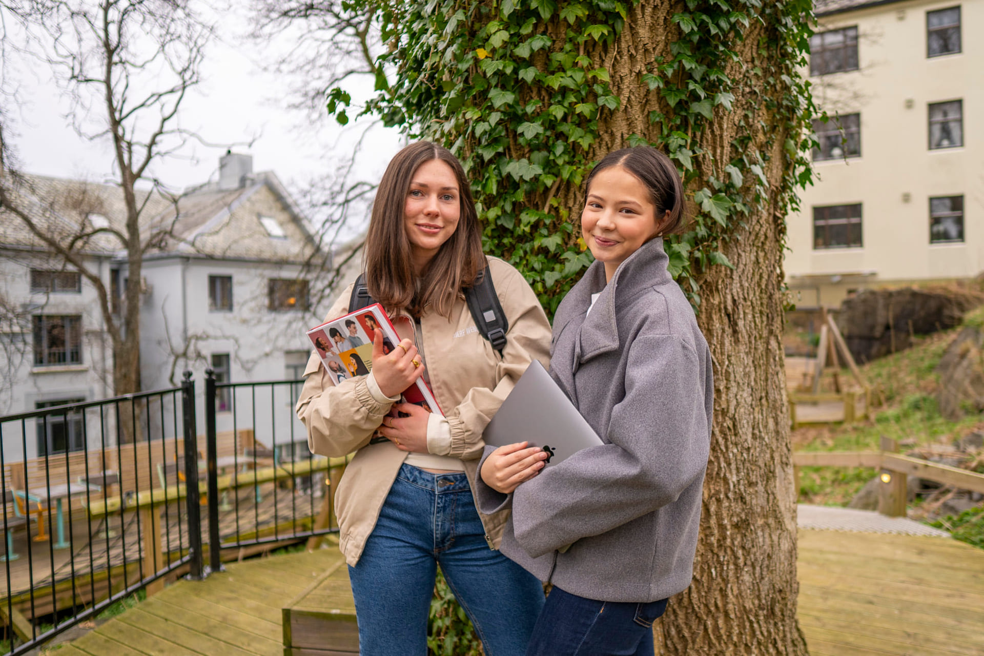 To privatister smiler til kamera mens de holder skolebok og macbook og står i en park