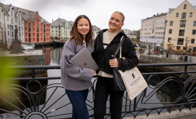 To jenter står ved siden av hverandre utendørs og har en macbook og et handlenett fra Akademiet