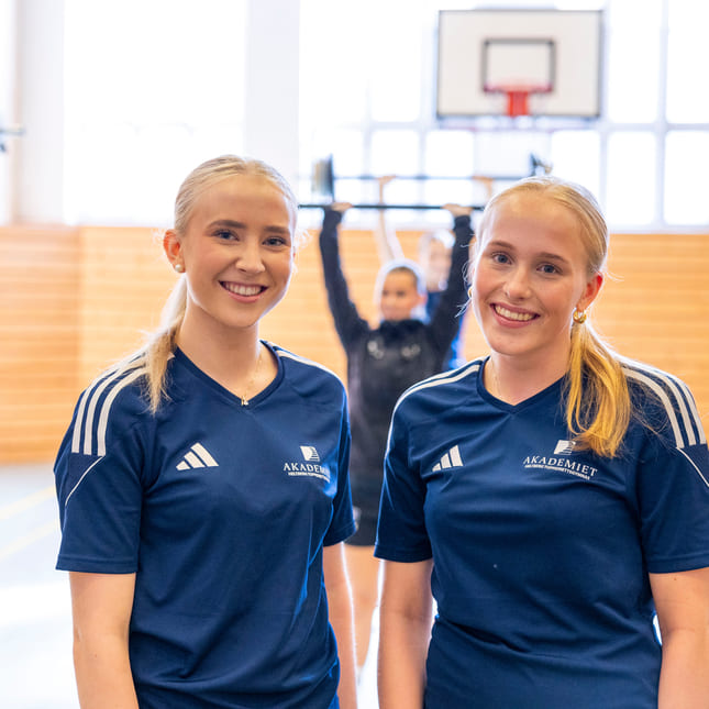 To elever smiler i en gymsal på Akademiet Ålesund.